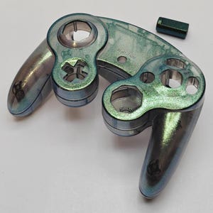 May include: A translucent game controller front shell with a shimmering green and blue glitter finish. The shell features cutouts for buttons, joysticks, and a D-pad, accompanied by a small, matching rectangular button component.