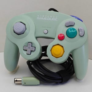 May include: A light green Nintendo GameCube controller featuring a gray analog stick, a D-pad, a yellow C-stick, a red button, a green button, gray X and Y buttons, a Start/Pause button, and a black wired cable with a light green connector plug.