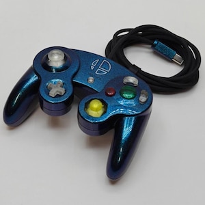Può includere: Un controller per videogiochi blu e nero con un cavo nero. Il controller ha una finitura metallica e presenta vari pulsanti colorati e un joystick. Il controller ha il logo Super Smash Bros.