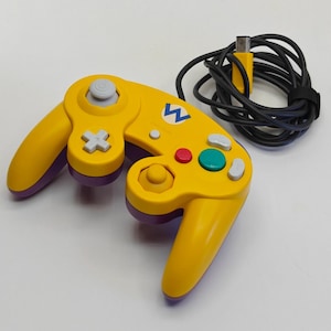 Peut inclure: Une manette de jeu vidéo jaune et violette avec un logo W bleu au centre. L'appareil comprend un joystick analogique gris, une croix directionnelle, un bouton rouge, un bouton vert et un câble noir avec une prise de connexion attachée.