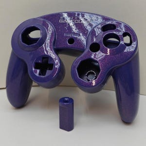 May include: A disassembled purple GameCube controller with a glittery finish. The controller has a D-pad, analog stick, and buttons. The text "NINTENDO GAMECUBE" is visible on the top. A separate purple component is also present.