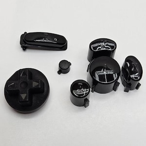 May include: A set of black replacement buttons for a video game controller. The buttons feature various symbols, including directional arrows, a cross, and letters.