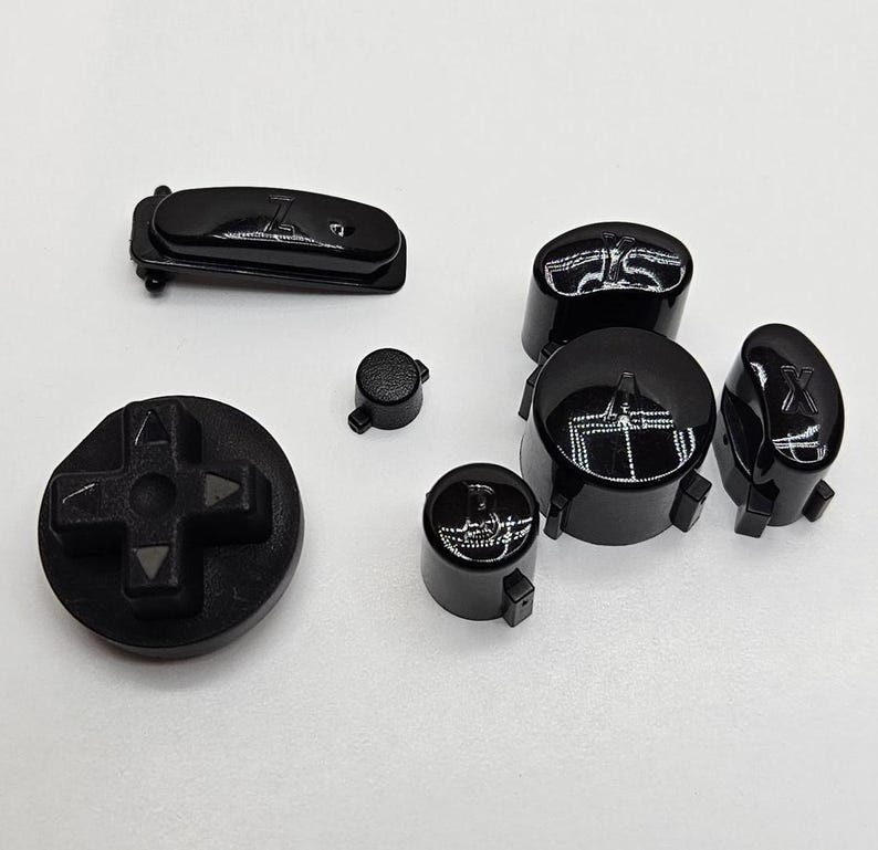 May include: A set of black replacement buttons for a video game controller. The buttons feature various symbols, including directional arrows, a cross, and letters.