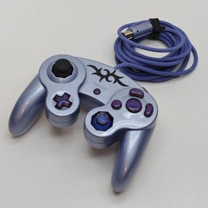 Peut inclure: Une manette de jeu vidéo bleu clair métallisé avec un motif tribal noir sur la face centrale. La manette comprend un joystick analogique noir, une croix directionnelle violette, des boutons d'action violets et un câble filaire bleu assorti.
