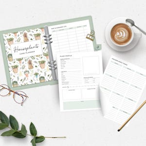 Peut inclure: Un planificateur de soins pour plantes d'int&eacute;rieur avec un classeur vert clair, ouvert pour r&eacute;v&eacute;ler des pages lign&eacute;es. La couverture pr&eacute;sente un motif aquarelle de plantes en pot. Une tasse de caf&eacute;, des lunettes et un crayon sont &eacute;galement sur la table.