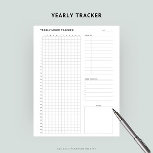 May include: A white Yearly Mood Tracker planner with a grid for daily entries, color key, mood boosters, and notes sections. The text "YEARLY TRACKER" is at the top. A silver pen rests on the bottom right corner. The Etsy shop name "AZILESCO PLANNERS ON ETSY" is at the bottom.