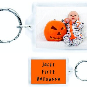 以下が含まれることがあります： ハロウィンをテーマにしたオレンジとブラックのパーソナライズされたキーホルダー。赤ちゃんがハロウィンコスチュームを着てジャック・オ・ランタンを持っている写真が写っています。キーホルダーには「Jacks First Halloween」という文字が書かれています。
