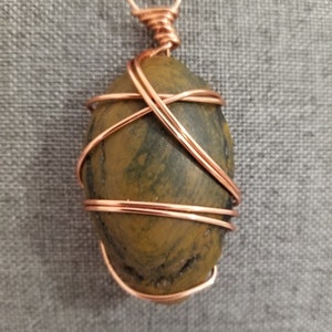 May include: A copper wire wrapped pendant featuring a green and brown stone. The stone is oval shaped and has a natural, rough texture.