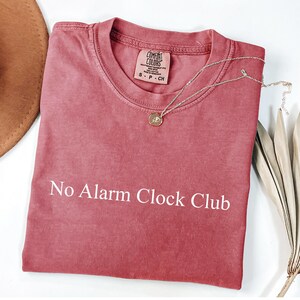 May include: A dusty rose-colored t-shirt with the text "No Alarm Clock Club" in white. A gold-colored pendant necklace rests on the shirt. The shirt is folded and has a Comfort Colors tag. A brown hat and dried plant are in the background.
