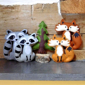 May include: A set of felt animal toys, including two raccoons, a fox, and a tree. The toys are made of soft, plush felt and have embroidered details. The raccoons are gray with black stripes, the fox is orange with white accents, and the tree is green with a brown trunk.