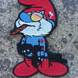May include: Embroidered patch featuring a cartoon character with a red hat, blue shirt, and red shoes. The character is holding a cigar in its mouth and has a mischievous expression.
