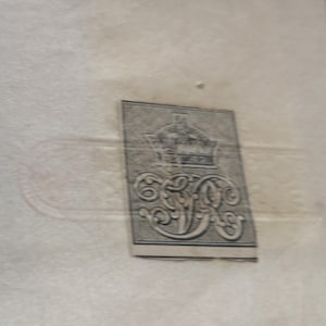 May include: A rectangular, grey and black label featuring a crown and elaborate initials. The label is positioned at an angle against a pale beige backdrop. The design is intricate, suggesting an antique or vintage origin.