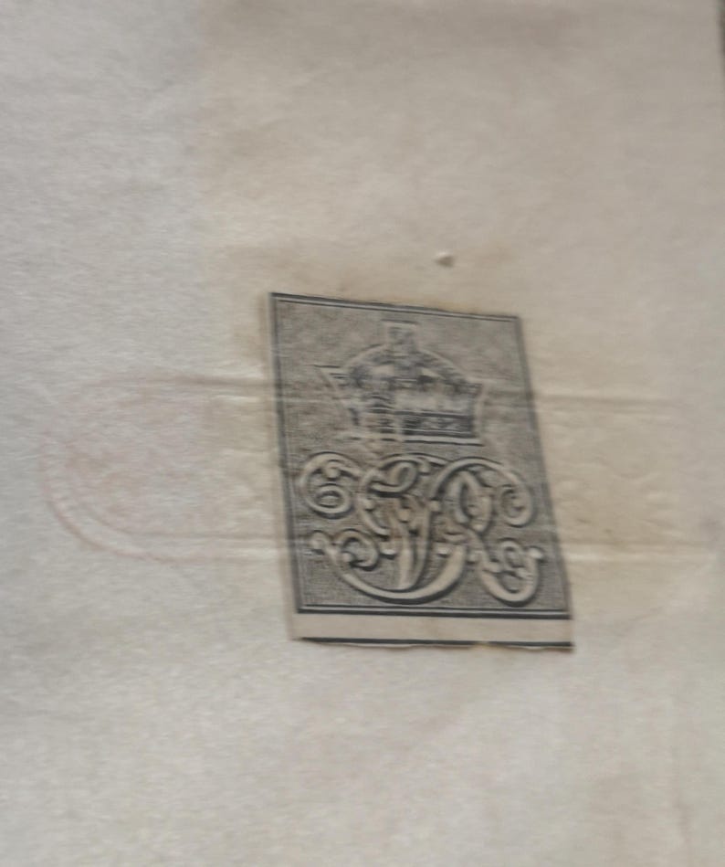 May include: A rectangular, grey and black label featuring a crown and elaborate initials. The label is positioned at an angle against a pale beige backdrop. The design is intricate, suggesting an antique or vintage origin.