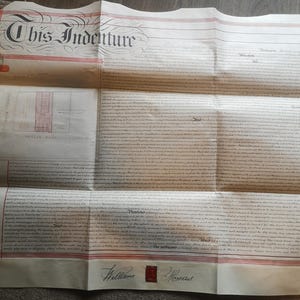 May include: An antique document titled "This Indenture" with ornate calligraphy. The paper is aged, with red stripes and text throughout. A small map and signatures are visible. The document is folded.