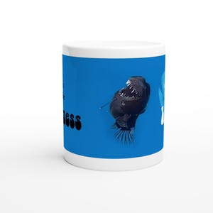 Op de afbeelding: Een witte keramische mok met een blauwe achtergrond waarop een zwarte anglerfish met een grote bek en een bioluminescerend lokmiddel is afgebeeld. De tekst "Embrace the Darkness" is in zwart op de mok gedrukt.