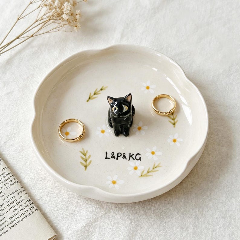 May include: A cream-colored ceramic ring dish with a scalloped edge, featuring a black cat figurine, daisy and leaf accents, and the text "L&P&KG". Two gold rings are placed on the dish.