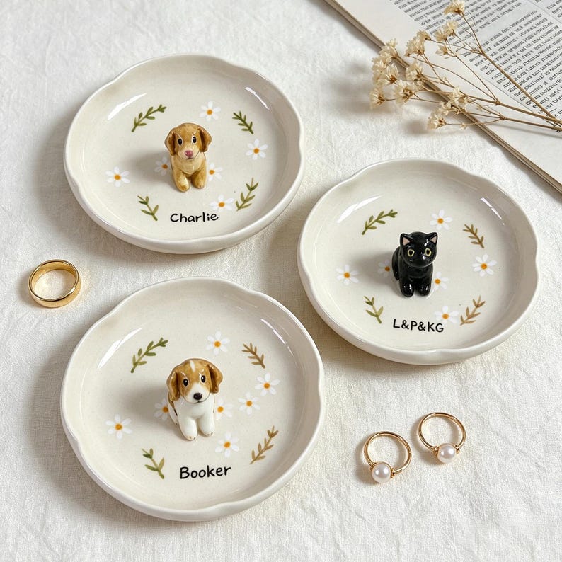 May include: Three ceramic trinket dishes with scalloped edges. Each dish features a small animal figurine: a dog, a cat, and another dog. The dishes are decorated with daisy and leaf motifs. The names "Charlie" and "Booker" are printed on two of the dishes, and "L&P&KG" on the third.
