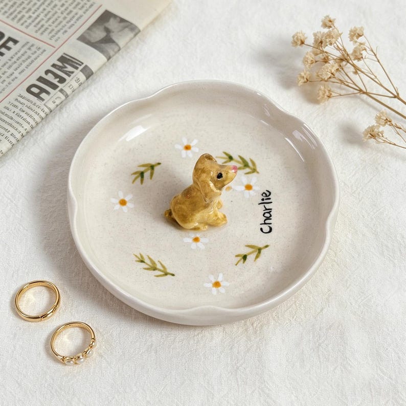 May include: A beige ceramic trinket dish with a scalloped edge, featuring a small, golden-colored dog figurine. The dish is decorated with painted daisies, green leaves, and the name "Charlie". Two gold rings are visible.