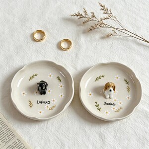 May include: Two small, off-white ceramic ring dishes with scalloped edges. One features a black cat figurine and the text "L&P&KG", the other a puppy figurine and the name "Booker". Both are decorated with daisies and green leaves.