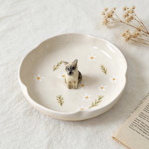 May include: A cream-colored ceramic dish with a scalloped edge, adorned with white daisies and green leaves. A small, detailed figurine of a cat with gray and tan markings sits in the center. The dish is approximately 8 inches in diameter.