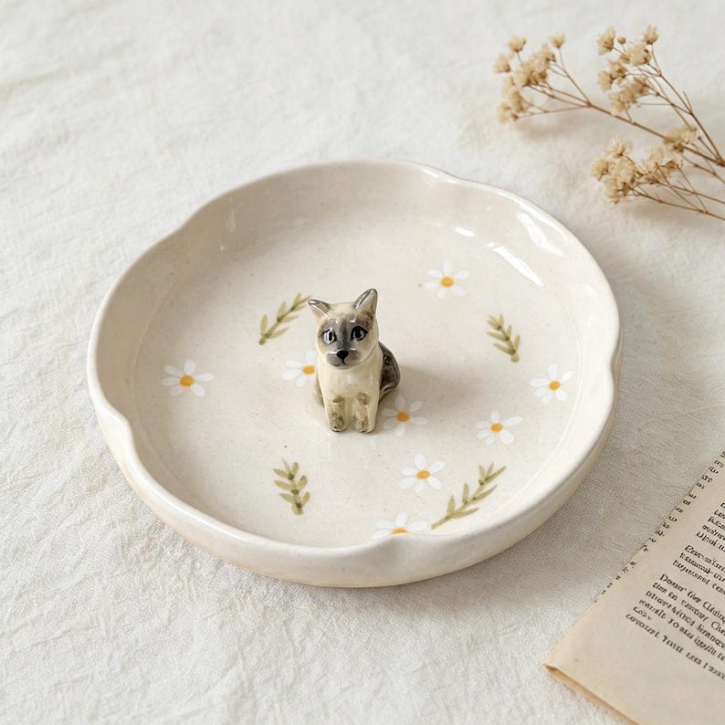 May include: A cream-colored ceramic dish with a scalloped edge, adorned with white daisies and green leaves. A small, detailed figurine of a cat with gray and tan markings sits in the center. The dish is approximately 8 inches in diameter.
