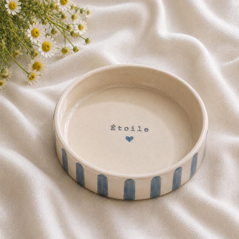 K&ouml;nnte beinhalten: Eine runde, cremefarbene Keramikschale mit vertikalen blauen Streifen an der Au&szlig;enseite. Der innere Boden zeigt das Wort &Eacute;toile in schwarzer Schrift &uuml;ber einem kleinen blauen Herzsymbol. Die Schale hat eine glatte, gl&auml;nzende Oberfl&auml;che.