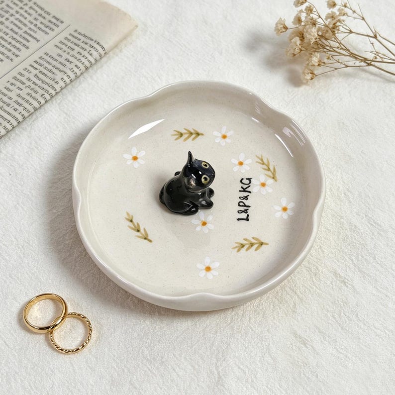 May include: A small, off-white ceramic dish with a scalloped edge, featuring a black cat figurine in the center. The dish is decorated with white daisy and green leaf designs, and the text "L&P&KG". Two gold rings are in the foreground.