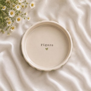K&ouml;nnte beinhalten: Eine runde, cremefarbene Keramikschale mit glattem Innenraum, auf dem der Name Figaro in schwarzer Schrift &uuml;ber einem kleinen, gr&uuml;nen Herzsymbol aufgedruckt ist. Die Schale hat einen gewellten Rand und liegt neben wei&szlig;en G&auml;nsebl&uuml;mchen.