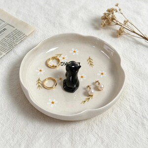 May include: A cream-colored ceramic trinket dish with a scalloped edge, featuring a black penguin figurine and daisy and leaf accents. The dish holds two gold rings and a pair of pearl earrings. A decorative and functional item for storing jewelry.