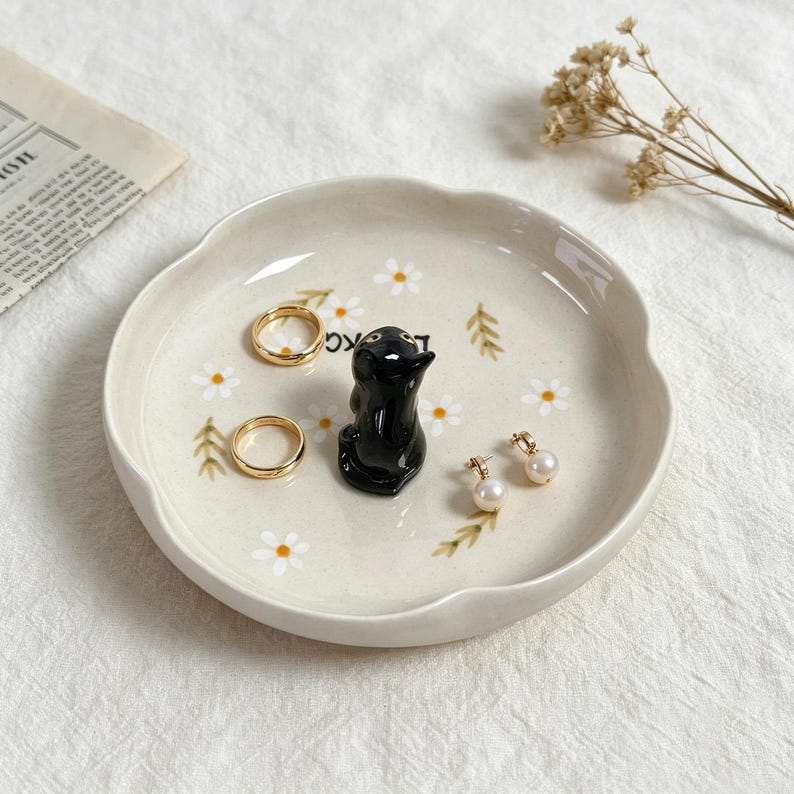 May include: A cream-colored ceramic trinket dish with a scalloped edge, featuring a black penguin figurine and daisy and leaf accents. The dish holds two gold rings and a pair of pearl earrings. A decorative and functional item for storing jewelry.