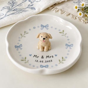 May include: A white ceramic trinket dish with a scalloped edge, featuring a central 3D figurine of a light brown puppy. The dish is decorated with a blue painted wreath of bows, leaves, and dots, with the text Mr & Mrs and 12.05.2025 printed.