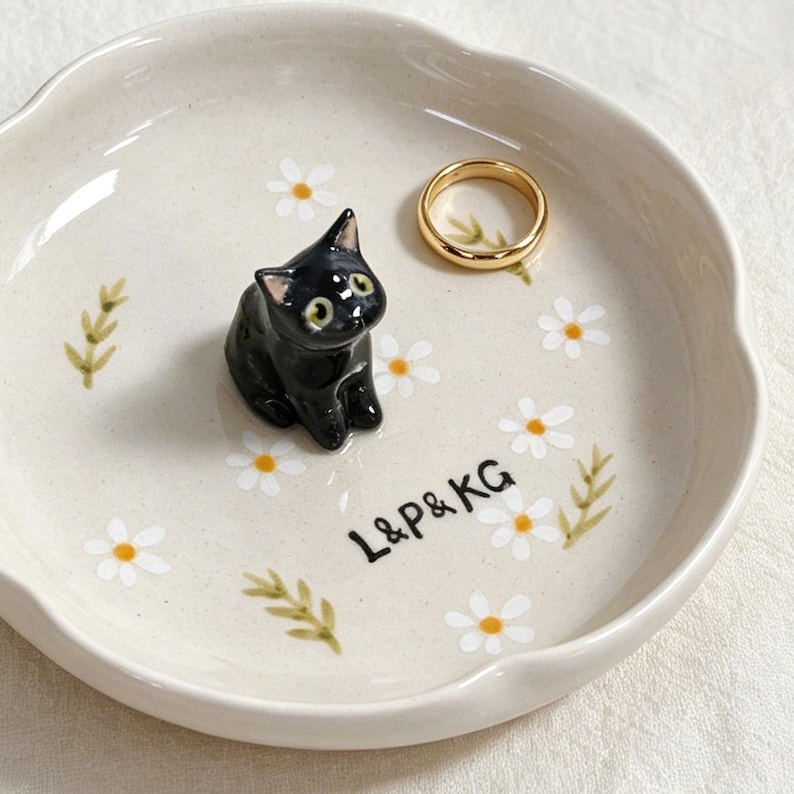 May include: A cream-colored ceramic ring dish with a scalloped edge, adorned with painted white daisies and green leaves. A small black cat figurine sits on the dish, next to a gold ring. The text "L&P&KG" is also on the dish.