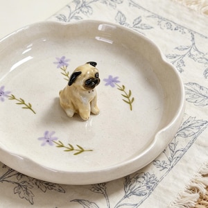 May include: A beige ceramic dish with a scalloped edge, decorated with lavender flowers and green stems. A small pug figurine with black ears and a black snout sits in the center of the dish. The dish is on a floral patterned cloth.