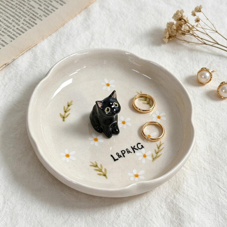 May include: A cream-colored ceramic ring dish with a black cat figurine in the center. The dish is decorated with white daisies and green leaves. Two gold rings and a pair of pearl earrings are also present. The text "L&P&KG" is printed on the dish.