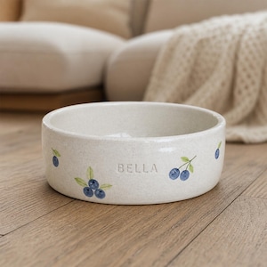 Peut inclure: Un bol rond en céramique blanc cassé avec une texture mouchetée, orné de grappes de myrtilles bleues peintes avec des feuilles vertes sur l'extérieur. Le nom BELLA est gravé en lettres capitales sur la face avant du bol.
