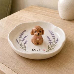 Peut inclure: Un vide-poche rond en céramique blanc cassé à bord ondulé, orné d'une petite figurine de chien brun aux poils bouclés au centre. Le plat est décoré de brins de lavande violets et du texte Mochi accompagné d'un petit cœur rose.