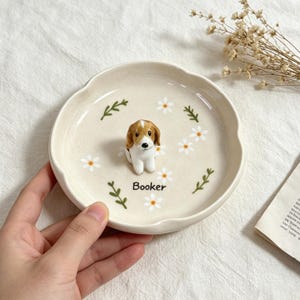May include: A cream-colored ceramic dish with a scalloped edge, featuring a small, detailed beagle figurine in the center. The dish is decorated with white daisy flowers and green sprigs. The name "Booker" is printed on the dish.