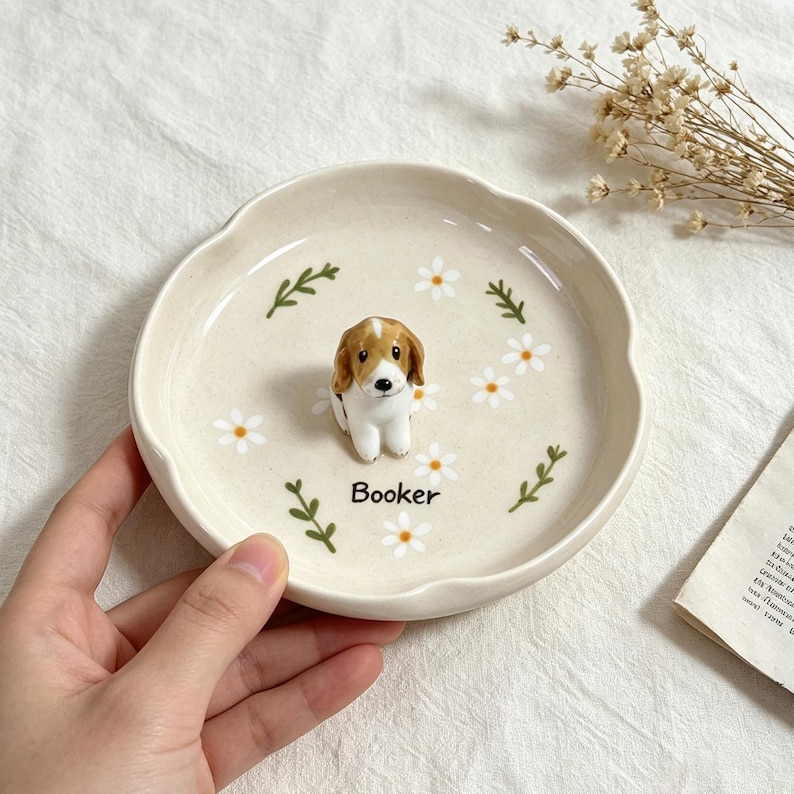 May include: A cream-colored ceramic dish with a scalloped edge, featuring a small, detailed beagle figurine in the center. The dish is decorated with white daisy flowers and green sprigs. The name "Booker" is printed on the dish.