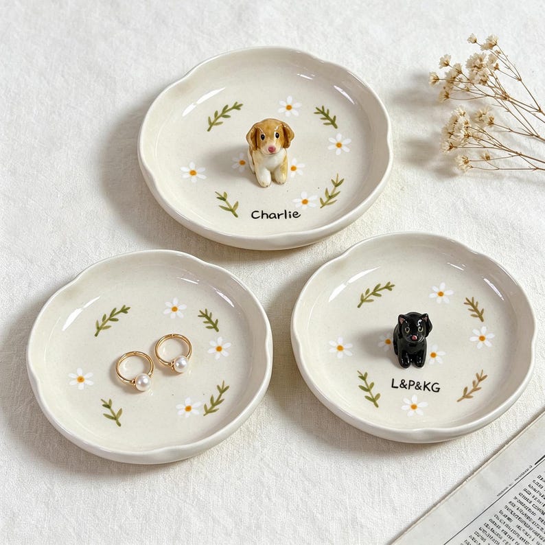 May include: Three off-white ceramic trinket dishes with scalloped edges. Each dish features a floral design with a small animal figurine in the center. One dish has a dog figurine and the name "Charlie". Another has two rings. The last has a black cat figurine and the text "L&P&KG".