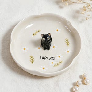 May include: A ceramic dish with a scalloped edge, featuring a black cat figurine in the center. The dish is decorated with white daisies, green leaves, and the text "L&P&KG". The overall color scheme is cream, black, white, and green.