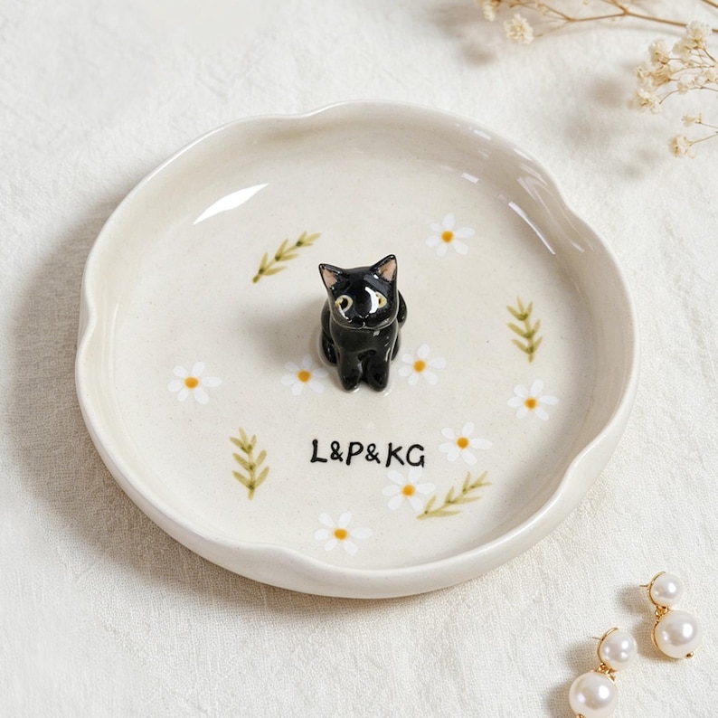 May include: A ceramic dish with a scalloped edge, featuring a black cat figurine in the center. The dish is decorated with white daisies, green leaves, and the text "L&P&KG". The overall color scheme is cream, black, white, and green.
