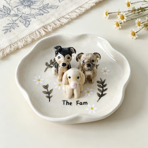 May include: A cream-colored ceramic dish with a scalloped edge, featuring three small dog figurines. The dish is decorated with painted daisies and the text "The Fam". The dogs are black and white, tan, and cream-colored.
