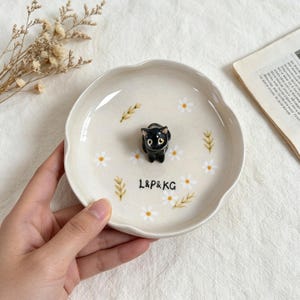 May include: A ceramic trinket dish with a scalloped edge, featuring a black cat figurine in the center. The dish is decorated with white daisies and green wheat stalks. The text "L&P&KG" is printed on the dish.