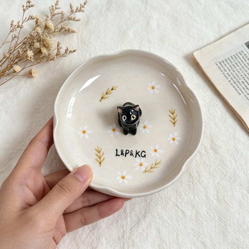 May include: A ceramic trinket dish with a scalloped edge, featuring a black cat figurine in the center. The dish is decorated with white daisies and green wheat stalks. The text "L&P&KG" is printed on the dish.