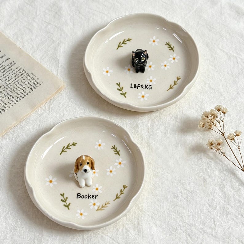 May include: Two off-white ceramic dishes with scalloped edges. One features a black cat figurine, the other a beagle figurine. Both are decorated with white daisies and green leaves. The first dish has the text "L&P&KG", and the second dish has the name "Booker".