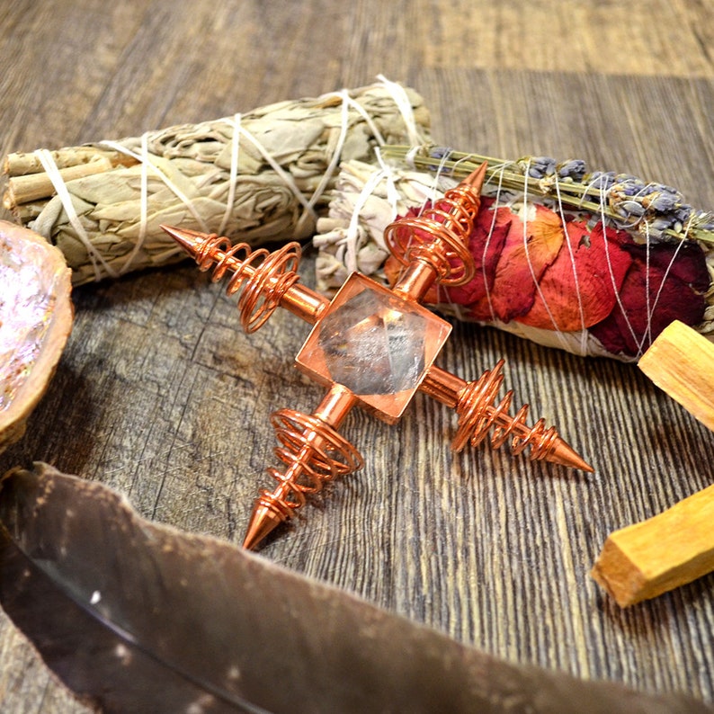 May include: A copper crystal grid with a clear quartz crystal in the center. The grid has four points with copper wire spirals. The grid is surrounded by sage bundles, dried flowers, and a wooden stick.