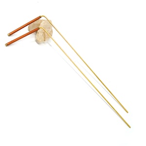May include: Two gold metal hair sticks with brown wooden tips. The sticks are lying on a white surface.