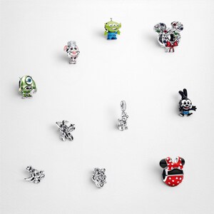 May include: A collection of ten silver-toned metal charms featuring various animated character designs, including a green alien, a one-eyed monster, a red polka-dot bow, a black and white rabbit, and other detailed miniature figures with enamel accents.