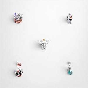 May include: A collection of five silver charms featuring various characters. Included are a pumpkin-themed charm with a purple hat, a snowman figure, a small elephant with large ears, a holiday-themed mouse charm, and a mermaid-inspired charm.