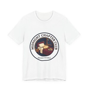 Op de afbeelding: Wit T-shirt met een cirkelvormig ontwerp. Het ontwerp bevat de tekst "MIDNIGHT CHAPTER CLUB" en "lifetime member." De afbeelding toont een slaapkamer met een bed, boeken, een kat en een nachtkastje.
